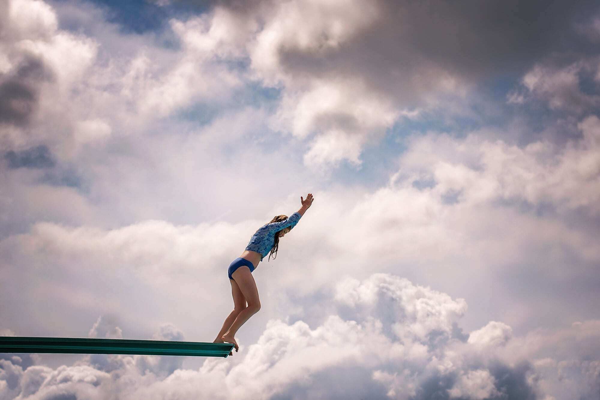 girl in diving position on diving board with blue sky and clouds in background 